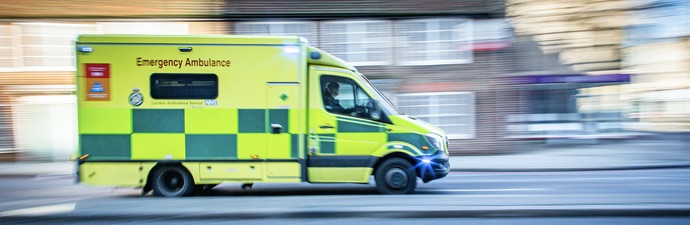 Yellow and green ambulance speeding through a suburban street in the UK.
