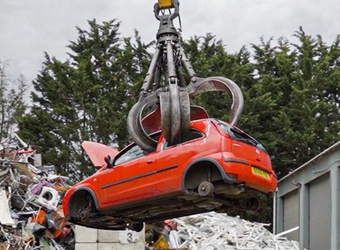 Red car being crushed and lifted by a grabber in a scrap metal yard.